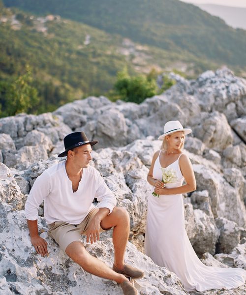 Bride with a bouquet stands on a rock next to sitting groom and looks away. High quality photo