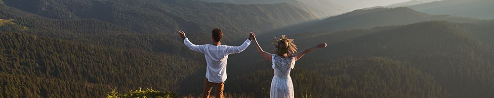 Back view of happy couple in white clothes is standing on top of mountain. Woman and boyfriend are holding hands and admiring mountains. Concept of beautiful landscape.